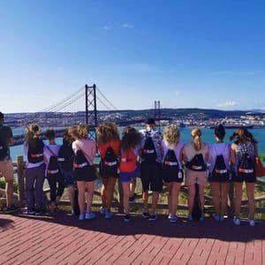 Eine WeRoad-Gruppe steht an einem Aussichtspunkt und blickt auf eine große rote Hängebrücke und eine Stadt jenseits des Wassers unter klarem blauem Himmel.