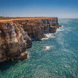 Imponentes acantilados de roca en capas se extienden hacia un reluciente océano azul bajo un cielo despejado.