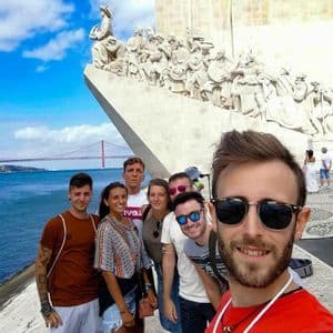 Eine WeRoad-Gruppe macht ein Selfie vor einem großen Steinmonument mit Brücke und Wasser im Hintergrund.