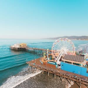 An aerial view of an amusement park pier with a large Ferris wheel over the ocean, next to a sandy beach under a clear sky.