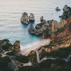 Eine Luftaufnahme einer Holztreppe, die sich einen grünen, felsigen Hang hinunter zu einem abgelegenen Sandstrand mit großen Felsnadeln im Wasser windet.