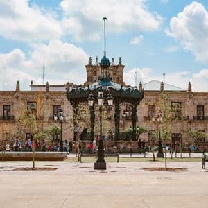 Un quiosco ornamentado y una farola antigua en una gran plaza pública frente a un edificio histórico de piedra bajo un cielo nublado.