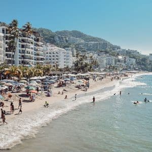 Gente relajándose bajo sombrillas y nadando en el mar en una playa soleada con hoteles en la ladera.