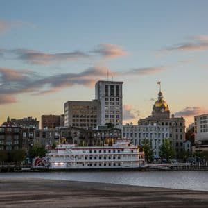 The Georgia Queen riverboat is docked on a river in front of a city skyline with a golden dome building at sunset.