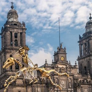Una enorme marioneta de esqueleto con sombrero monta un caballo esqueleto frente a una catedral de piedra ornamentada con dos campanarios.