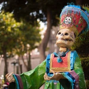 Una gran figura de esqueleto decorada, con un tocado colorido y un traje vibrante, se encuentra al aire libre frente a frondosos árboles verdes.