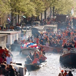 Una grande folla di persone festeggia su barche durante un festival lungo un trafficato canale cittadino fiancheggiato da case galleggianti.