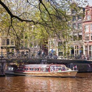 Un battello turistico naviga sull'acqua sotto un ponte con pedoni e biciclette, con alte case sui canali sullo sfondo.