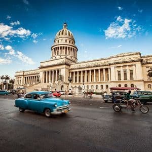 Auto d'epoca colorate sfilano su una strada davanti a un grande edificio classico con una cupola sotto un cielo blu.