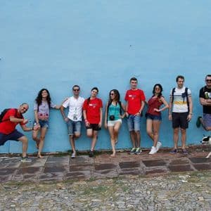 Un viaje en grupo de WeRoad posa para una foto contra una pared azul brillante en una calle empedrada, con dos perros pequeños cerca.