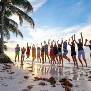 Un viaggio di gruppo WeRoad che salta insieme su una spiaggia tropicale al tramonto, con palme lungo la riva.
