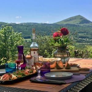 Une table dressée avec vin, fromage et salade sur une terrasse avec vue panoramique sur des collines verdoyantes et une montagne.