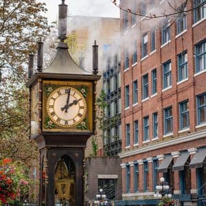An ornate steam clock releases steam from its pipes on a city street next to a red brick building.