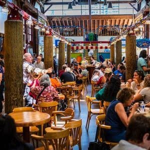 A bustling indoor food hall filled with people eating at wooden tables and walking around under a high ceiling with wooden beams.