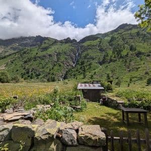 Un chalet rustique dans une prairie de montagne verdoyante avec une cascade lointaine, vu par-dessus un mur de pierre au premier plan.