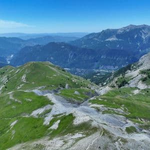 Vue aérienne d'une vaste chaîne de montagnes avec des collines verdoyantes et des pentes rocheuses sous un ciel bleu clair.