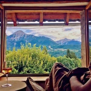Une personne se détend avec un verre de vin devant une fenêtre ouverte, contemplant une vallée verdoyante et une chaîne de montagnes.
