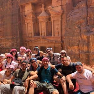 A WeRoad group trip smiles for a photo in front of a large temple carved into a sunlit, reddish rock face.