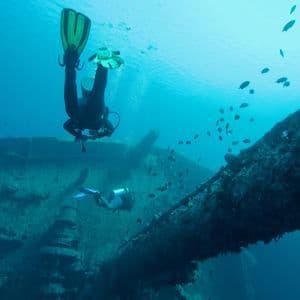 Two divers from a WeRoad group trip explore a sunken shipwreck underwater, with schools of small fish swimming by.