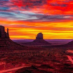 Three large rock buttes are silhouetted against a vibrant red and orange sunset over a desert landscape with a winding road.