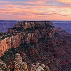 A panoramic view of a vast, layered canyon with a prominent mesa under a colorful pink and orange sunset sky.