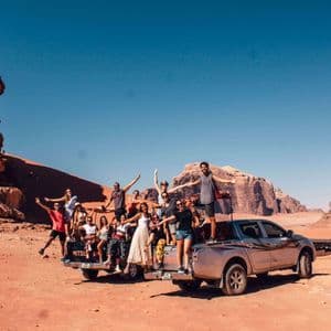 Un voyage de groupe WeRoad avec des personnes souriantes posant sur un pick-up dans un désert aux formations rocheuses rouges.
