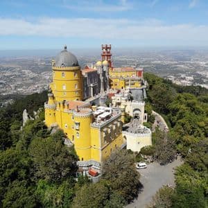 Un château coloré jaune et rouge avec des tours se dresse sur une colline boisée surplombant une ville tentaculaire sous un ciel bleu.