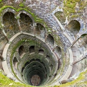 Vue plongeante sur un puits en pierre moussue, avec un escalier en colimaçon descendant le long de ses parois circulaires.