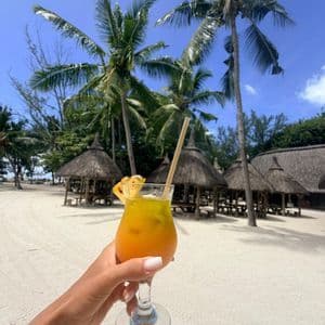 Une main tenant un cocktail orange sur une plage tropicale avec des palmiers et des cabanes au toit de chaume.