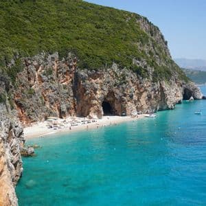 Eine Vogelperspektive auf einen abgelegenen Strand mit türkisfarbenem Wasser am Fuße einer großen, grün bewachsenen Klippe mit einer markanten Höhle.