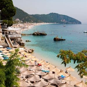 Ein belebter Strand mit Reihen von Strohschirmen säumt das Ufer einer türkisfarbenen Bucht, umgeben von grünen Hügeln.