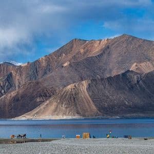 Un vasto lago blu si adagia ai piedi di imponenti montagne rocciose, con persone e animali sulla riva.