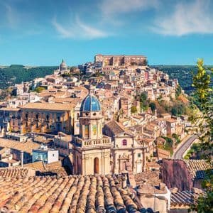 Veduta soleggiata dall'alto di una città storica costruita su un pendio, con tetti in terracotta e una cupola blu prominente.