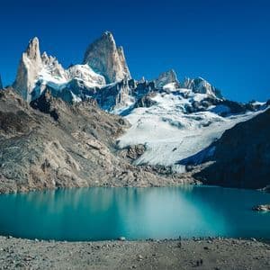 Un groupe WeRoad longe la rive rocheuse d'un lac turquoise, au pied de montagnes déchiquetées et enneigées, sous un ciel bleu clair.