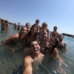 A man takes a selfie of his WeRoad group trip while they all swim and smile in the sea on a sunny day.