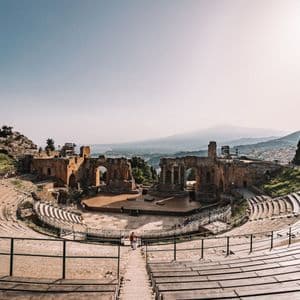 Ein Panoramablick auf ein antikes Steinamphitheater mit Bühne und Ruinen, das eine Küste und einen entfernten Berg unter klarem Himmel überblickt.