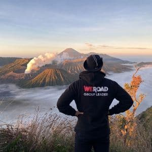 Un coordinador de grupo WeRoad visto de espaldas, contemplando volcanes humeantes que emergen de un mar de nubes al amanecer.