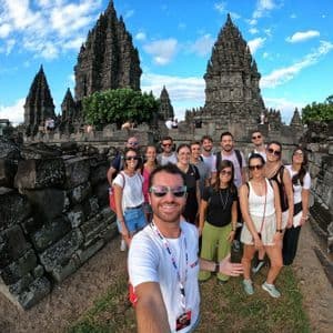 A WeRoad group trip takes a wide-angle selfie in front of an ancient stone temple complex under a partly cloudy sky.