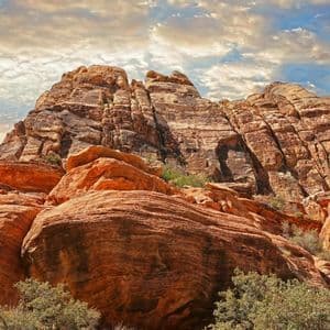 A large, layered red rock formation rises towards a partly cloudy sky, with green shrubs visible at its base.