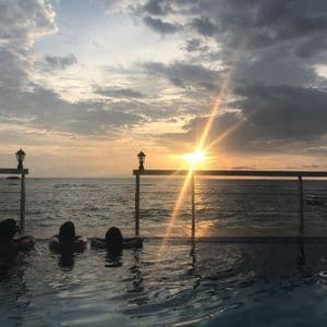Drei Personen einer WeRoad Gruppenreise entspannen in einem Infinity-Pool mit Blick auf das Meer während eines bewölkten Sonnenuntergangs.