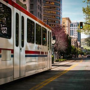A white UTA Trax tram on a track running down a tree-lined city street next to modern buildings.