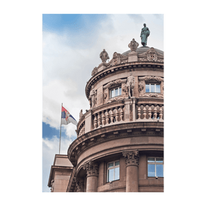 Inquadratura dal basso della bandiera serba che sventola accanto alla facciata ricurva e ornata di un edificio classico con statue.