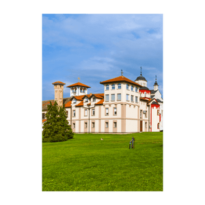 Un grande monastero con tetti di tegole rosse e cupole ortodosse si erge dietro un vasto prato verde sotto un cielo azzurro con nuvole leggere.
