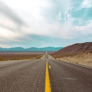 A long, straight asphalt road with a yellow line leads through a vast desert landscape toward distant mountains.
