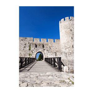 Un ponte di legno conduce all'ingresso ad arco di una fortezza in pietra con mura merlate e una torre, sotto un cielo azzurro e limpido.