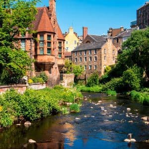 Une rivière traverse un village historique, ses rives bordées d'une végétation luxuriante et de vieilles bâtisses en pierre sous un ciel ensoleillé.