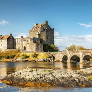 Un château de pierre est situé sur une petite île, relié par un pont de pierre voûté au-dessus de l'eau sous un ciel partiellement nuageux.