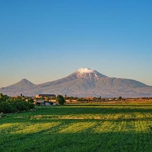 Une grande montagne enneigée se dresse au loin derrière un champ vert vif et un petit village à l'heure dorée.