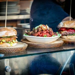Two burgers and a salad bowl with meat and vegetables are displayed on a stainless steel counter at a food stall.