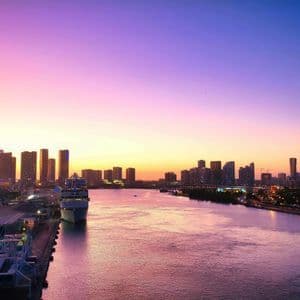 A city skyline along a wide waterway with a docked cruise ship, viewed under a purple and orange sunset.